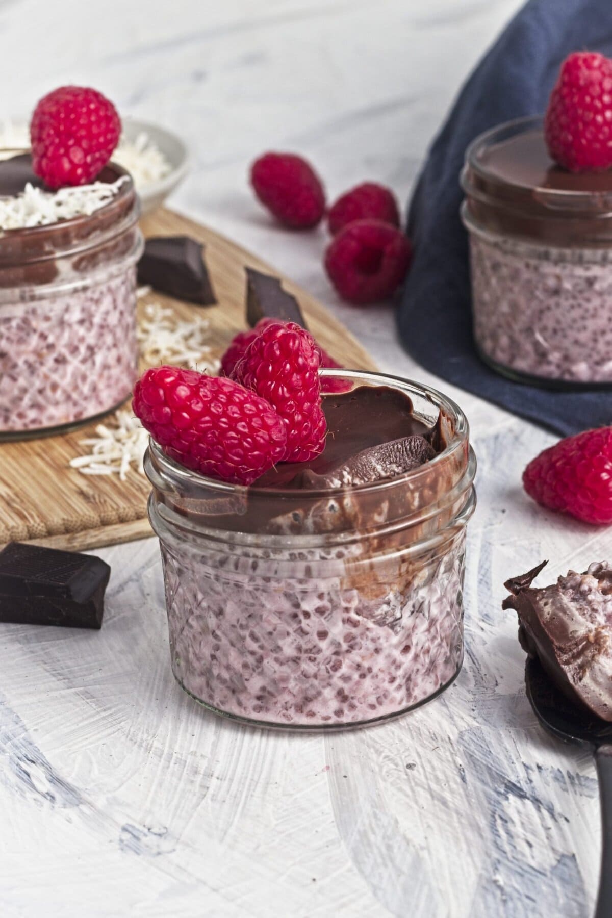 a wooden board with chocolate chunks, raspberries and coconut shreds with a bowl of shredded coconut and a jar of raspberry chia seed pudding with chocolate and coconut and a raspberry on top