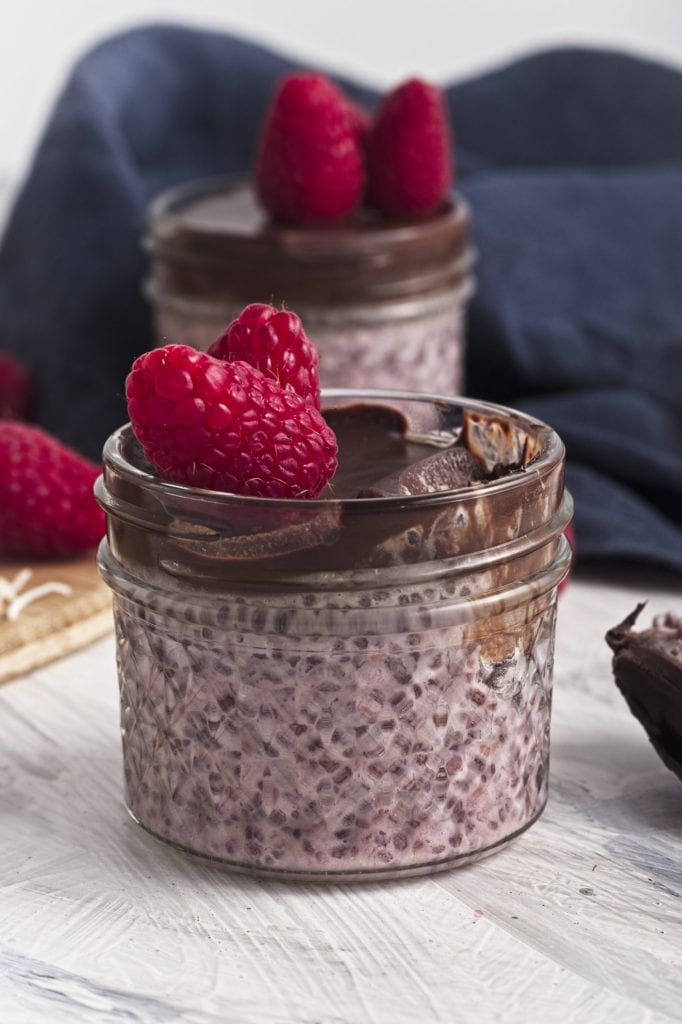 a wooden board with chocolate chunks, raspberries and coconut shreds with a bowl of shredded coconut and a jar of raspberry chia seed pudding with chocolate and coconut and a raspberry on top