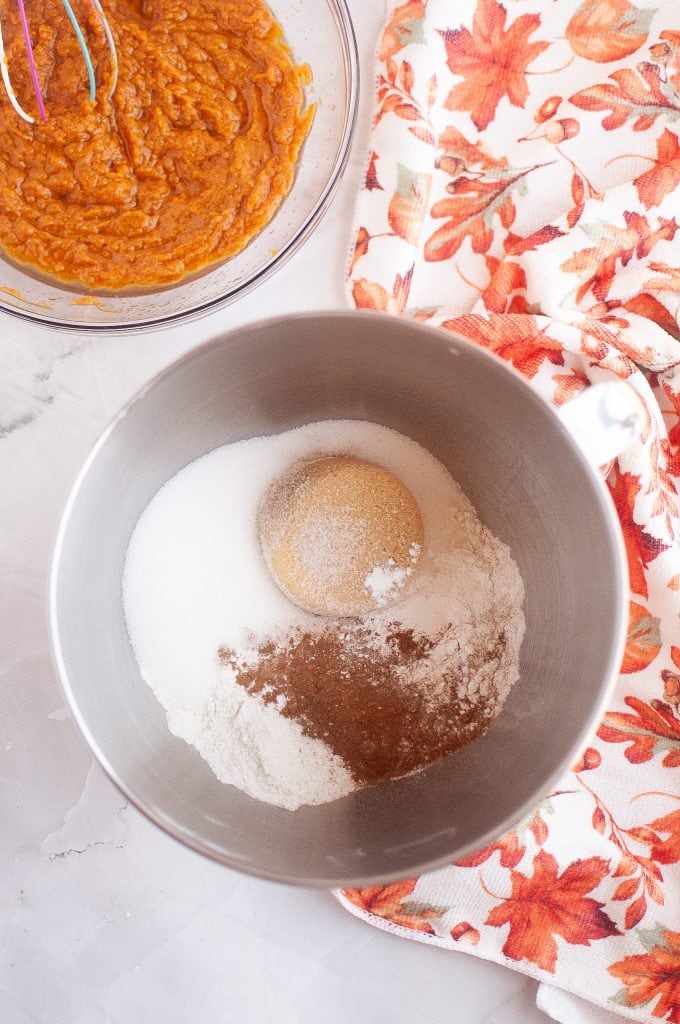 A metal bowl of dry ingredients for vegan pumpkin cookies in the center. The mixed bowl of wet ingredints in the top left corner with a fall napkin on the right.