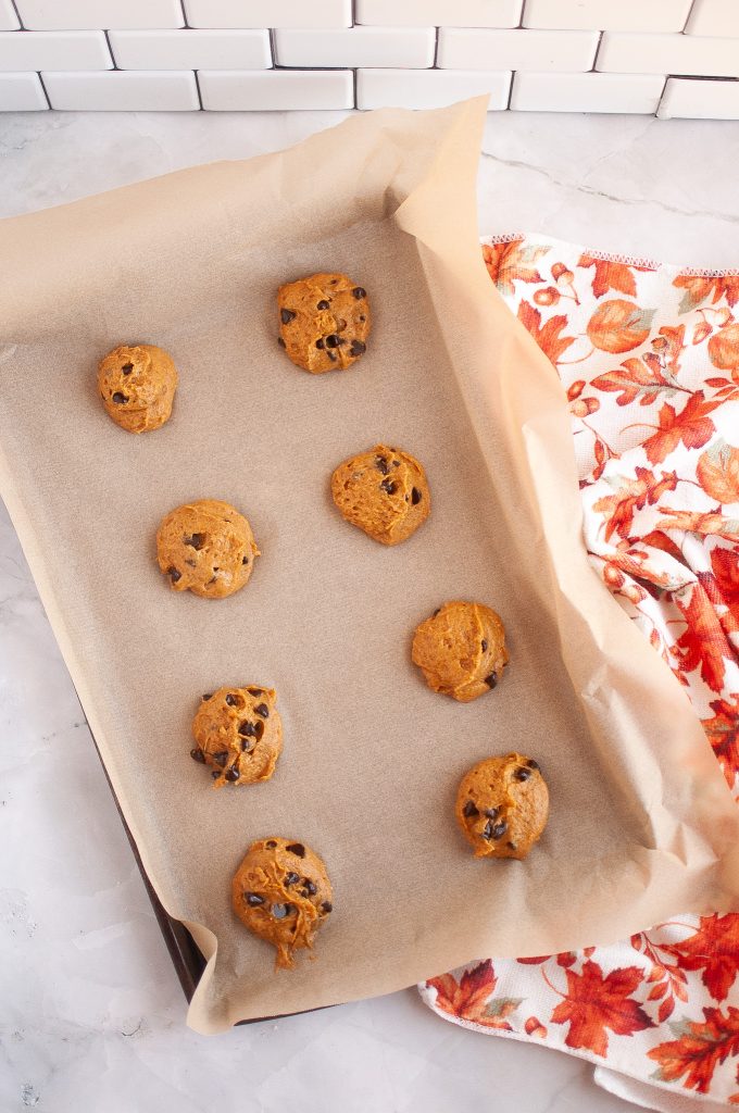 Eight vegan pumpkin chocoate chip cookies on a parchment -lined pan ready to bake. A fall-themed cloth is on the right.