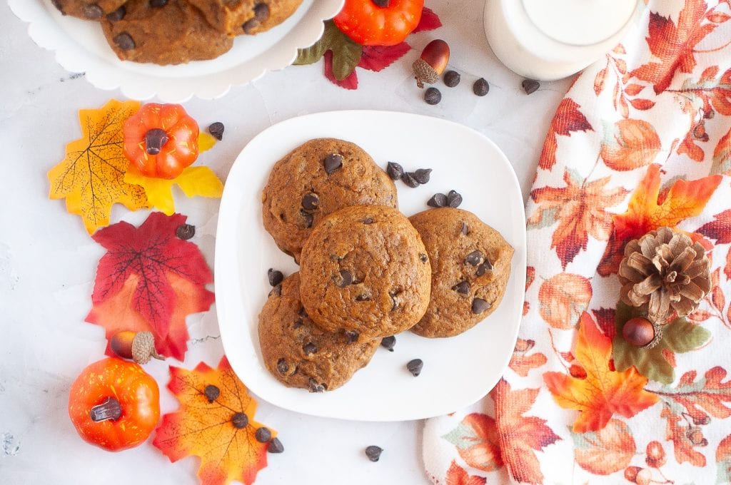 A square plate of four pumpkin cookies in the center with a plate of more in the top left corner. Almond milk in glasses top right corner. Cute fall decor surrounding