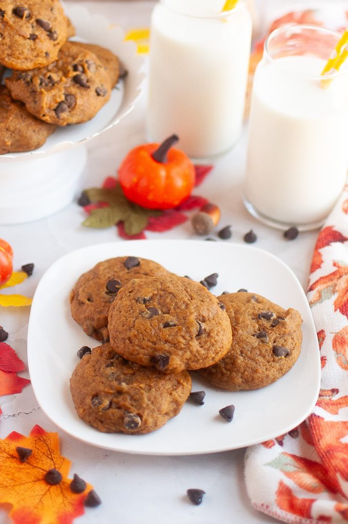 A square plate of four pumpkin cookies in the center with a plate of more in the top left corner. Almond milk in glasses top right corner. Cute fall decor surrounding