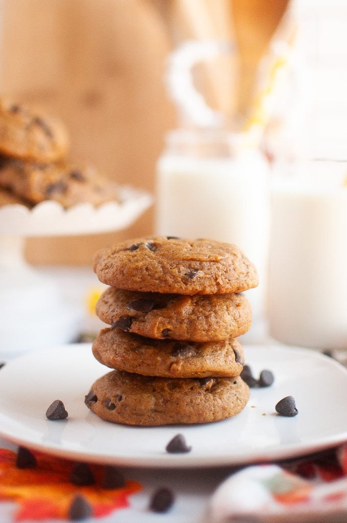 A square plate with a stack of four pumpkin cookies in the center with a plate of more in the top left corner. Almond milk in glasses top right corner. Cute fall decor surrounding