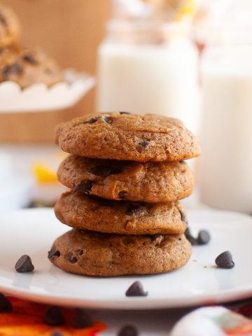 A square plate with a stack of four pumpkin cookies in the center with a plate of more in the top left corner. Almond milk in glasses top right corner. Cute fall decor surrounding