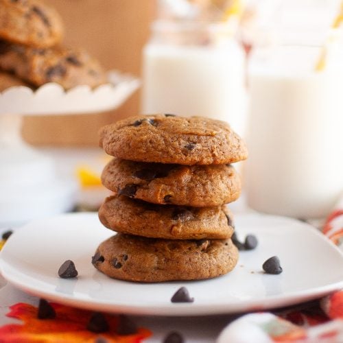 A square plate with a stack of four pumpkin cookies in the center with a plate of more in the top left corner. Almond milk in glasses top right corner. Cute fall decor surrounding