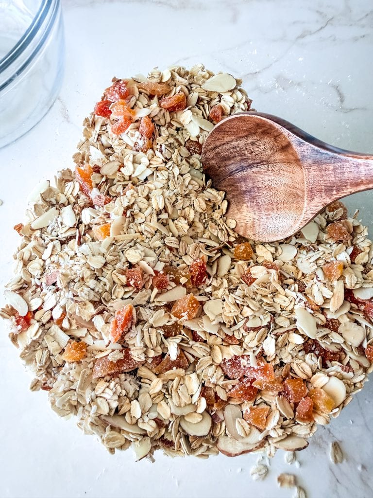 Golden oat and dried fruit granola mixture on a white surface with a wooden scoop, perfect for healthy vegan breakfast or snack.
