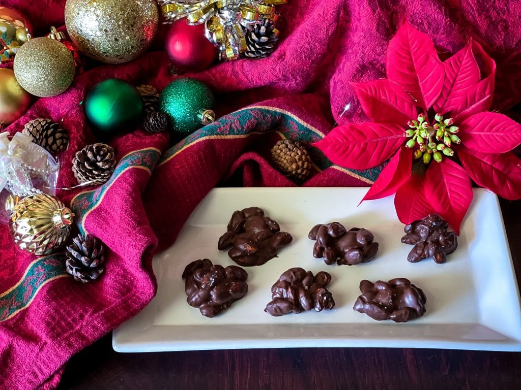 Chocolate almond clusters on a white plate with festive holiday decorations and red fabric background.