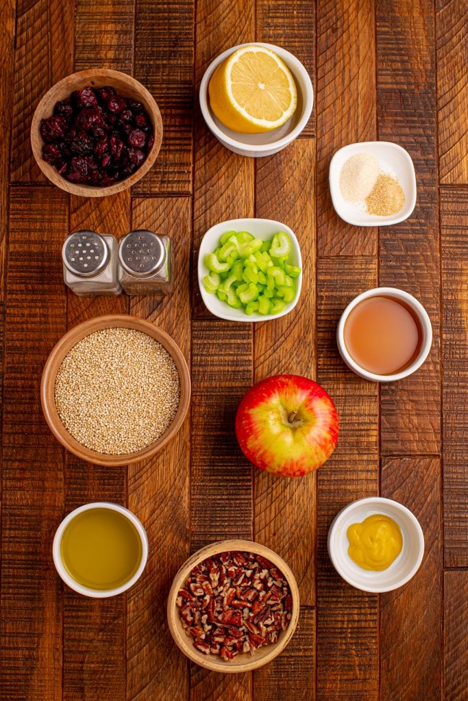 A flat lay of various food ingredients on a wooden surface, including an apple, lemon, celery, quinoa, pecans, dried cranberries, olive oil, mustard, agave, apple cider vinegar, spices, and salt and pepper shakers.