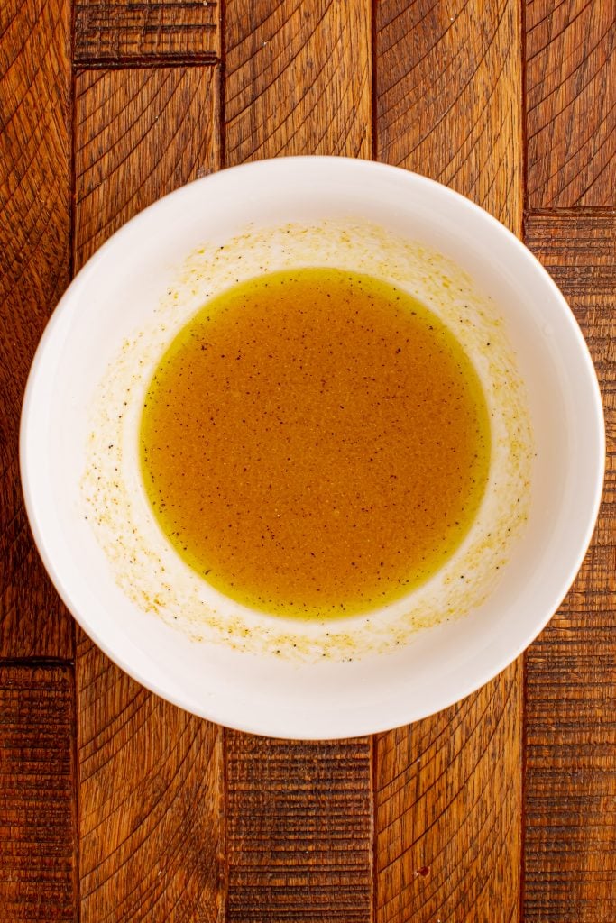 A white bowl filled with a golden-yellow vinaigrette dressing sits on a textured wooden surface. Perfect for drizzling over a fall quinoa salad, the dressing has a slightly oily appearance with visible seasoning specks.
