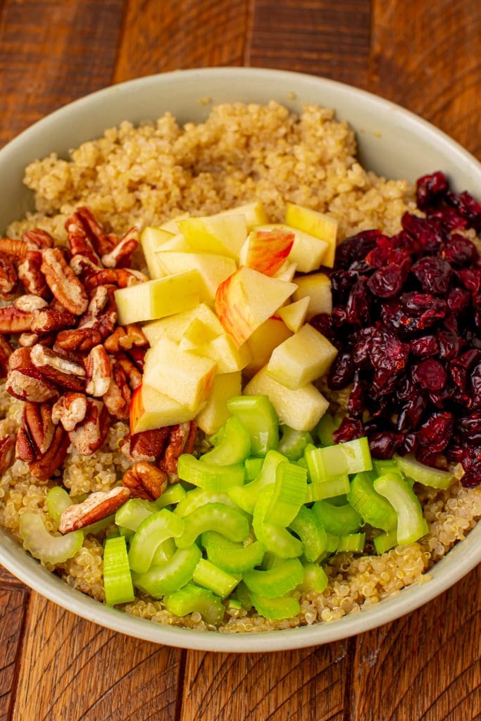 A bowl of quinoa salad with apples and cranberries, alongside celery slices and pecans, arranged in neat piles on a wooden table.