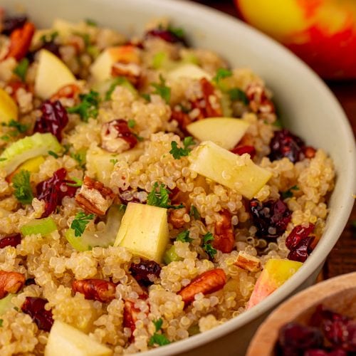 A bowl of quinoa salad with chopped apples, dried cranberries, celery, pecans, and fresh parsley, with whole apples and a small bowl of cranberries in the background.