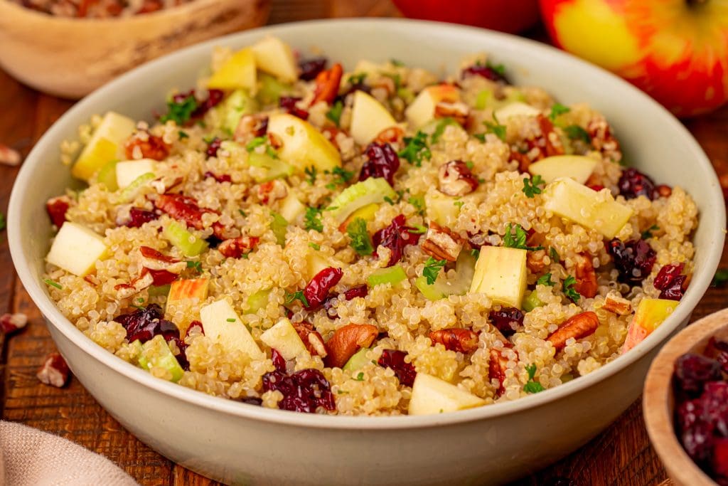 A bowl of quinoa salad with diced apples, celery, dried cranberries, chopped nuts, and fresh herbs, on a wooden table with apples and bowls of ingredients in the background.