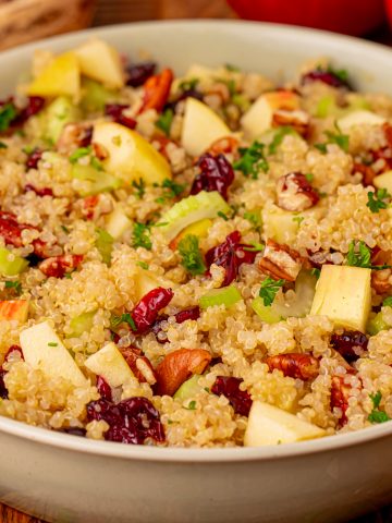 A bowl of quinoa salad with diced apples, celery, dried cranberries, chopped nuts, and fresh herbs, on a wooden table with apples and bowls of ingredients in the background.