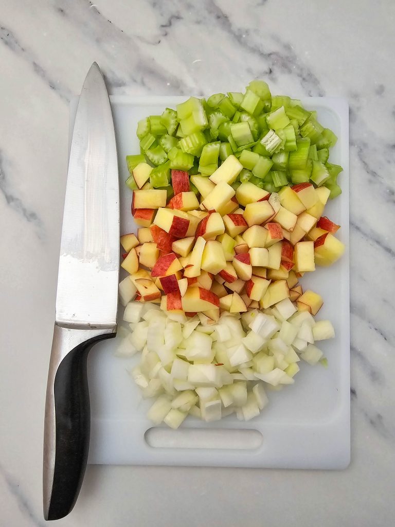Cutting board with knife on the left wide. Piles of chopped celery, apples, and onions.