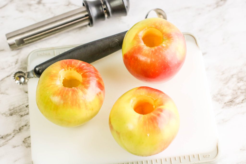 Three cored apples sit on a white cutting board next to a metal apple corer and a melon baller, all on a marble countertop.