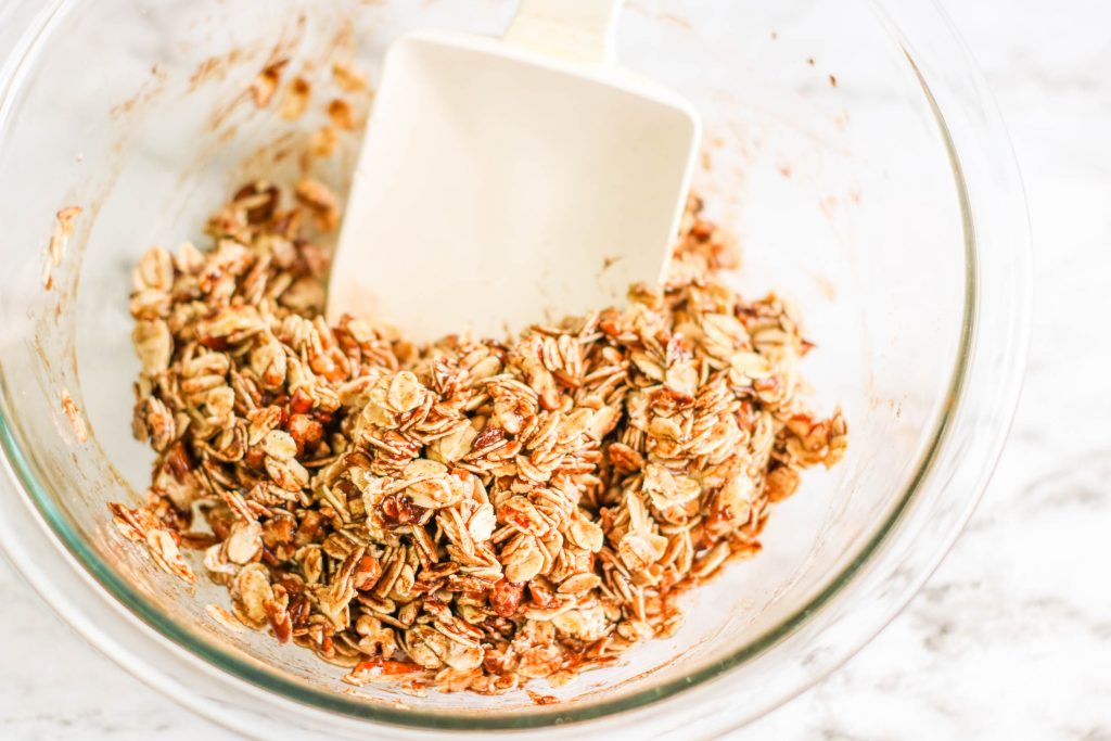 A glass bowl filled with a mixture of oats and spices, being stirred with a white spatula. The bowl sits on a white surface.