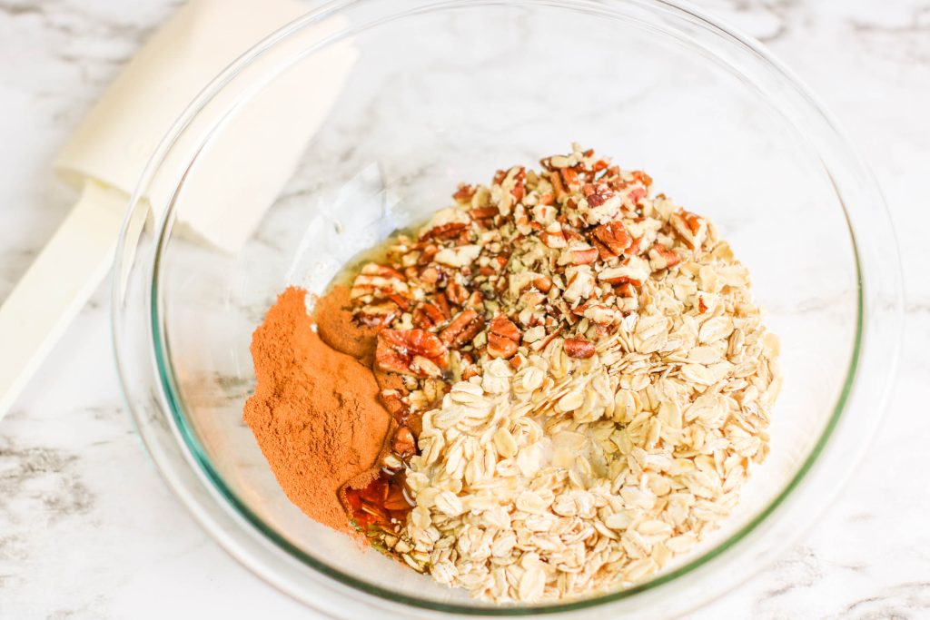 A glass bowl contains rolled oats, chopped pecans, ground cinnamon, and a drizzle of agave on a light countertop. A white spatula rests nearby.