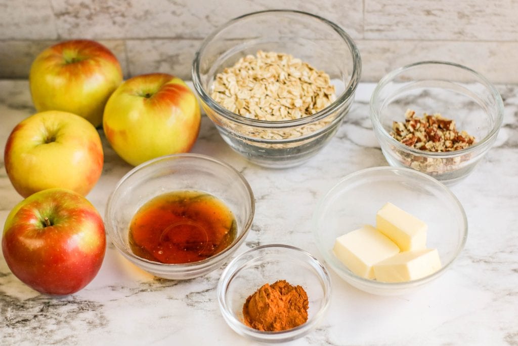 Five apples, a bowl of rolled oats, chopped pecans, vegan butter, maple syrup, and ground cinnamon are arranged on a white marble surface, with a tiled backsplash in the background.
