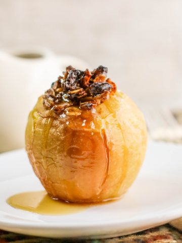 A baked apple filled with nuts and raisins sits on a white plate, drizzled with syrup. A fork, napkin, and white pitcher are in the blurred background.
