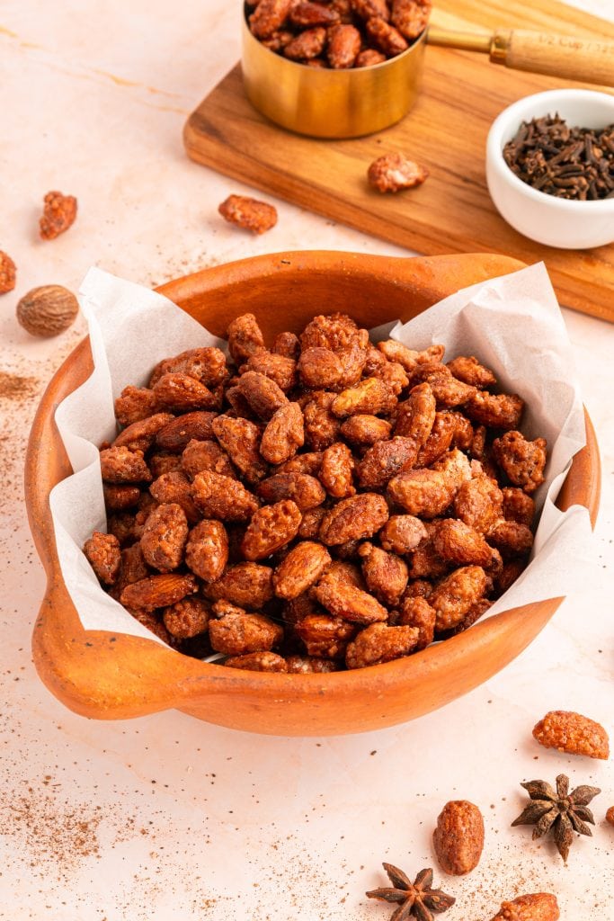 A wooden bowl lined with parchment paper is filled with sugar coated almonds. Some spices and loose almonds are scattered on the light-colored surface nearby, along with a small bowl and a wooden board in the background.