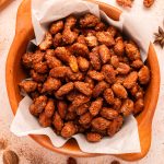 A wooden bowl lined with parchment paper, filled with roasted, caramelized almonds. Some almonds and star anise are scattered on a light-colored surface around the bowl.