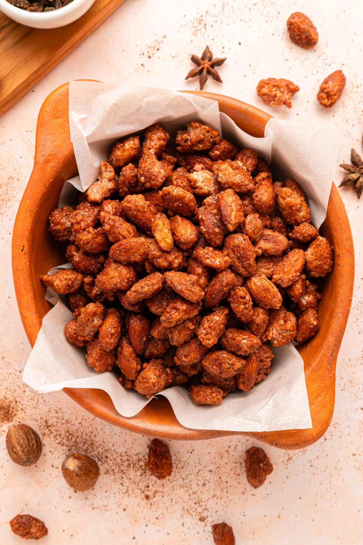 A wooden bowl lined with parchment paper, filled with roasted, caramelized almonds. Some almonds and star anise are scattered on a light-colored surface around the bowl.