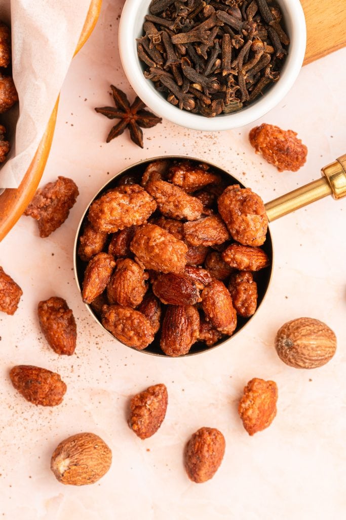 A brass measuring cup filled with candied almonds sits on a light surface, surrounded by whole spices including nutmeg, star anise, cloves in a white bowl, and scattered candied almonds.