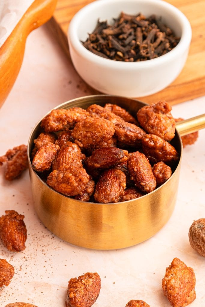 A brass measuring cup filled with candied nuts sits on a light surface, surrounded by scattered nuts. In the background, a small white bowl holds whole cloves.