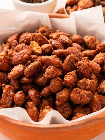 A brown ceramic bowl lined with parchment paper is filled with candied almonds. Some almonds are scattered on the light-colored surface nearby, and a small bowl of more almonds is visible in the background.