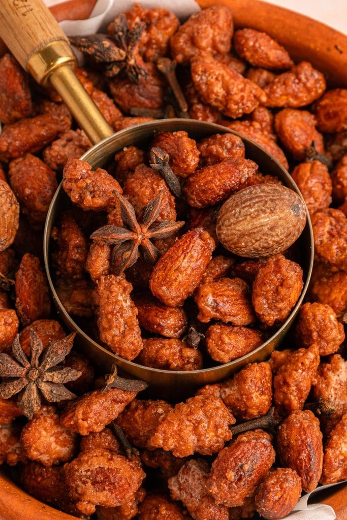 A bowl filled with candied almonds, whole star anise, and a whole nutmeg, with a metal scoop resting inside the bowl. The almonds are coated in a shiny, caramelized glaze.