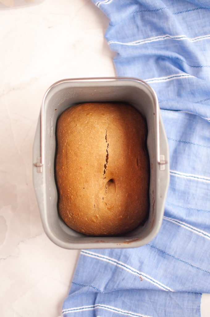 A loaf of peanut butter bread with a crack and small dent on top, freshly baked inside a bread machine pan, sits on a light countertop next to a blue cloth with white stripes.