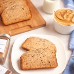 Two slices of yeasted peanut butter bread on a white plate, with a jar of peanut butter, a glass of milk, and spices nearby. More slices of banana bread are on a wooden board in the background. A blue cloth is to the right.
