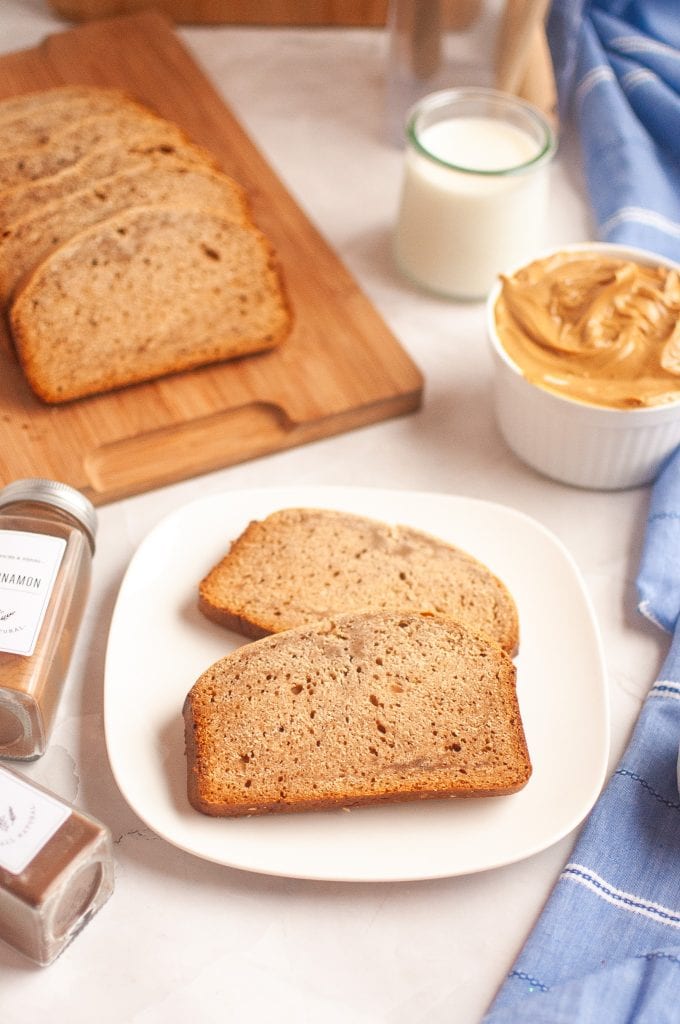 Two slices of yeasted peanut butter bread on a white plate, with a jar of peanut butter, a glass of milk, and spices nearby. More slices of banana bread are on a wooden board in the background. A blue cloth is to the right.