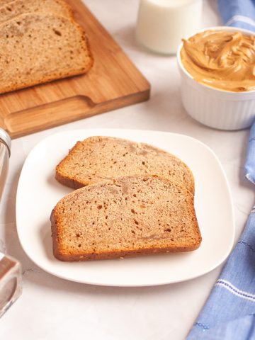 A plate with two slices of peanut butter bread sits beside jars of cinnamon and nutmeg, a small bowl of peanut powder with a spoon, a ramekin of peanut butter, a glass of milk, and a cutting board with more bread, all on a white surface.