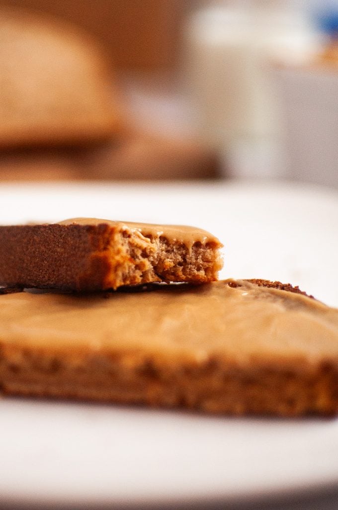 A close-up of a slice of peanut butter bread with a creamy peanut butter, topped with another slice that has a bite taken out of it, placed on a white surface with a blurred background.