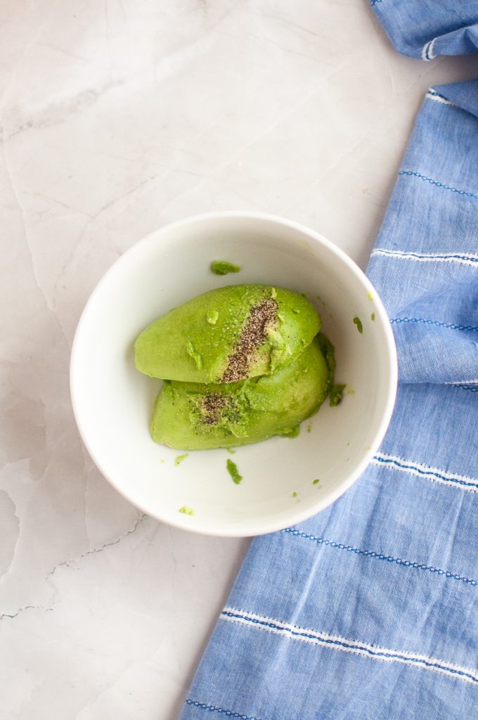 A white bowl with sliced avocado and ground black pepper inside, placed on a light marble surface next to a blue and white striped cloth.