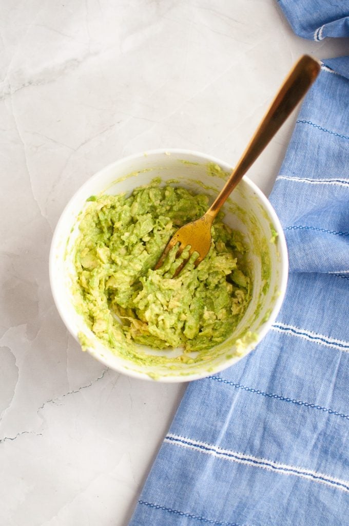 A bowl of mashed avocado with a gold fork in it, sitting on a marble surface next to a blue and white striped cloth.