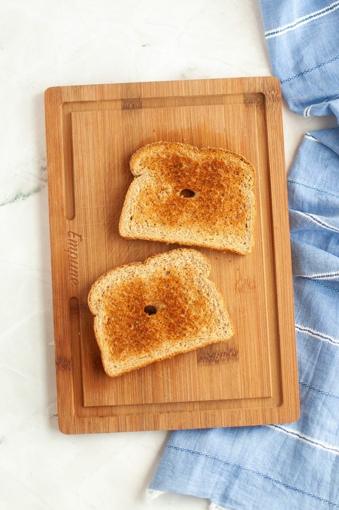 Two slices of toasted bread with holes in the center sit on a wooden cutting board next to a blue and white striped cloth, on a light-colored marble surface.