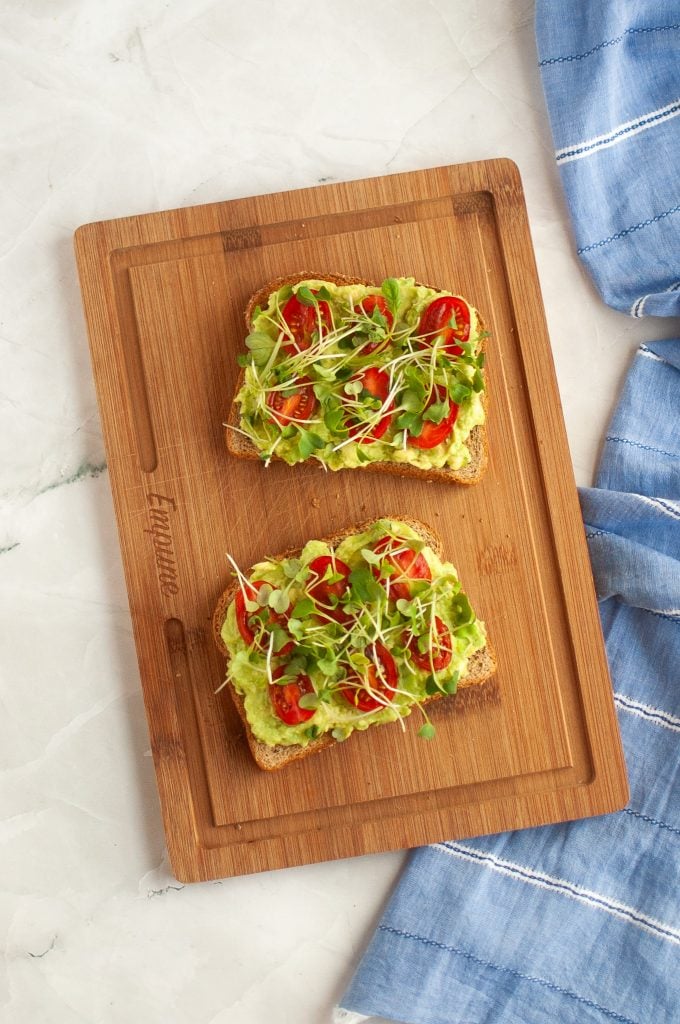 Two slices of toast topped with mashed avocado, tomato slices, lettuce, and microgreens are placed on a wooden cutting board. A blue cloth with white stripes lies nearby on a white marble surface.