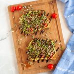 Two slices of vegan avocado toast topped with microgreens, seeds, and balsamic glaze are on a wooden board, garnished with cherry tomatoes. A blue striped cloth is placed to the right of the board.