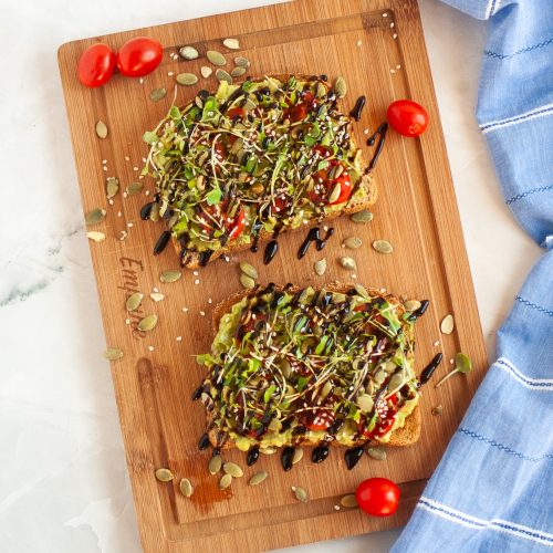 Two slices of vegan avocado toast topped with microgreens, seeds, and balsamic glaze are on a wooden board, garnished with cherry tomatoes. A blue striped cloth is placed to the right of the board.