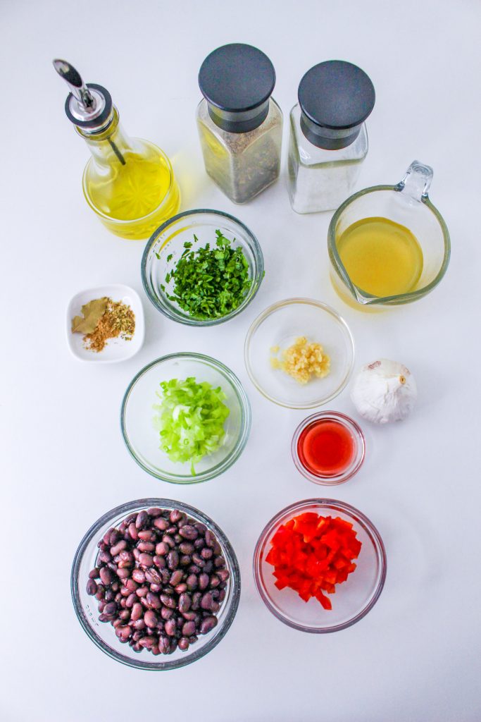 Overhead view of various ingredients for vegan cuban black beans in glass bowls on a white surface: black beans, diced red bell pepper, chopped green onions,, minced garlic, chopped herbs, dried spices, olive oil, vinegar, salt, pepper, and vegetable broth.