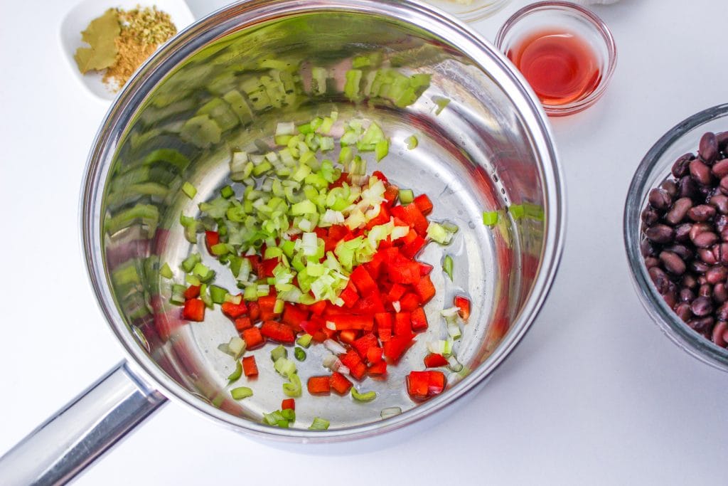 A metal pot with chopped celery and red bell pepper inside, surrounded by a bowl of black beans, a small glass bowl of red wine vinegar, and a plate with spices and bay leaves on a white surface.