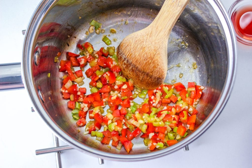 A stainless steel pot on a stove contains diced red bell peppers, green bell peppers, and chopped garlic being sautéed with a wooden spoon. A small glass cup with red wine vinegar is nearby.