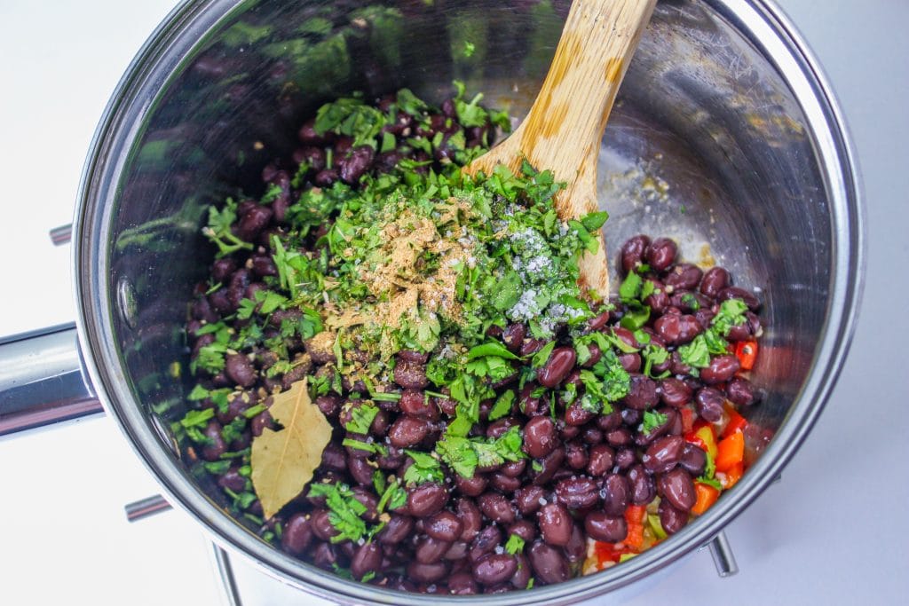 A pot filled with black beans, chopped cilantro, red bell peppers, seasonings, and a bay leaf, being mixed with a wooden spoon.