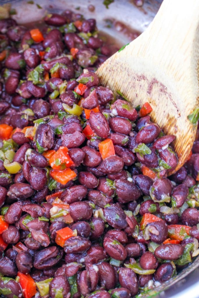 Close-up of cooked black beans mixed with chopped red and green bell peppers and herbs, being stirred with a wooden spoon.