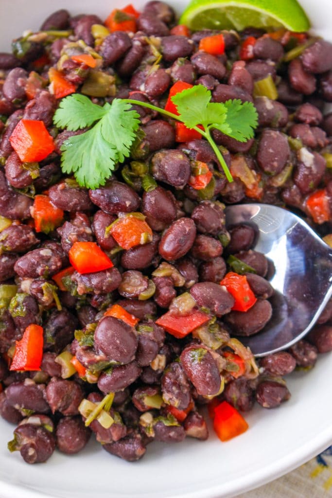 A bowl of Cuban black beans with chopped red bell peppers, green herbs, and a sprig of cilantro on top, garnished with lime wedges and served with a spoon.