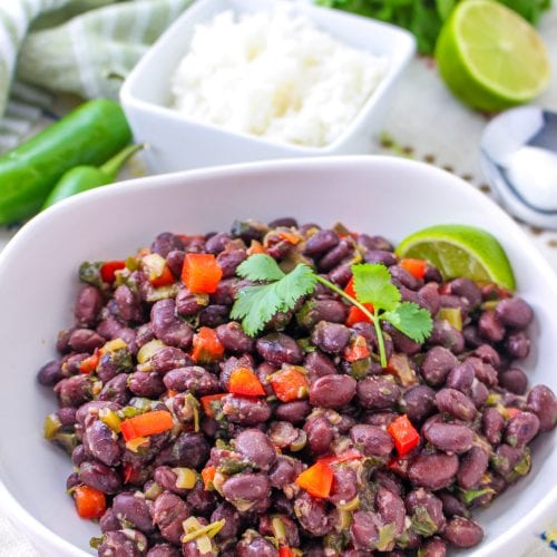 A white bowl filled with seasoned Cuban black beans, diced red peppers, and cilantro, garnished with a lime wedge. In the background are a dish of rice, lime halves, jalapeño, and fresh cilantro.