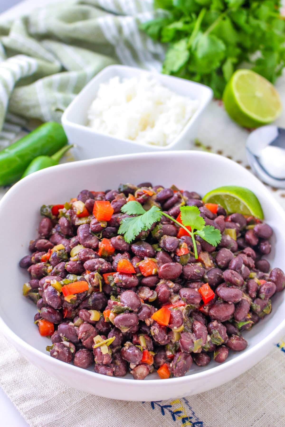 A white bowl filled with seasoned Cuban black beans, diced red peppers, and cilantro, garnished with a lime wedge. In the background are a dish of rice, lime halves, jalapeño, and fresh cilantro.