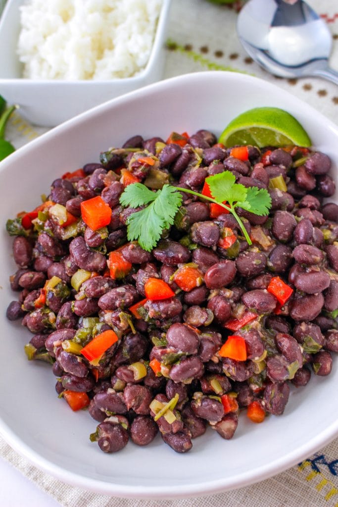 A bowl of seasoned Cubanblack beans mixed with chopped red bell peppers and herbs, garnished with fresh cilantro and a lime wedge, with a bowl of white rice and a spoon in the background.
